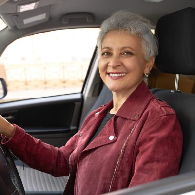 People, city and transportation concept. Portrait of cheerful attractive 60 year old woman with short hairstyle smiling broadly at camera with hand on steering wheel, sitting in driver seat in new car